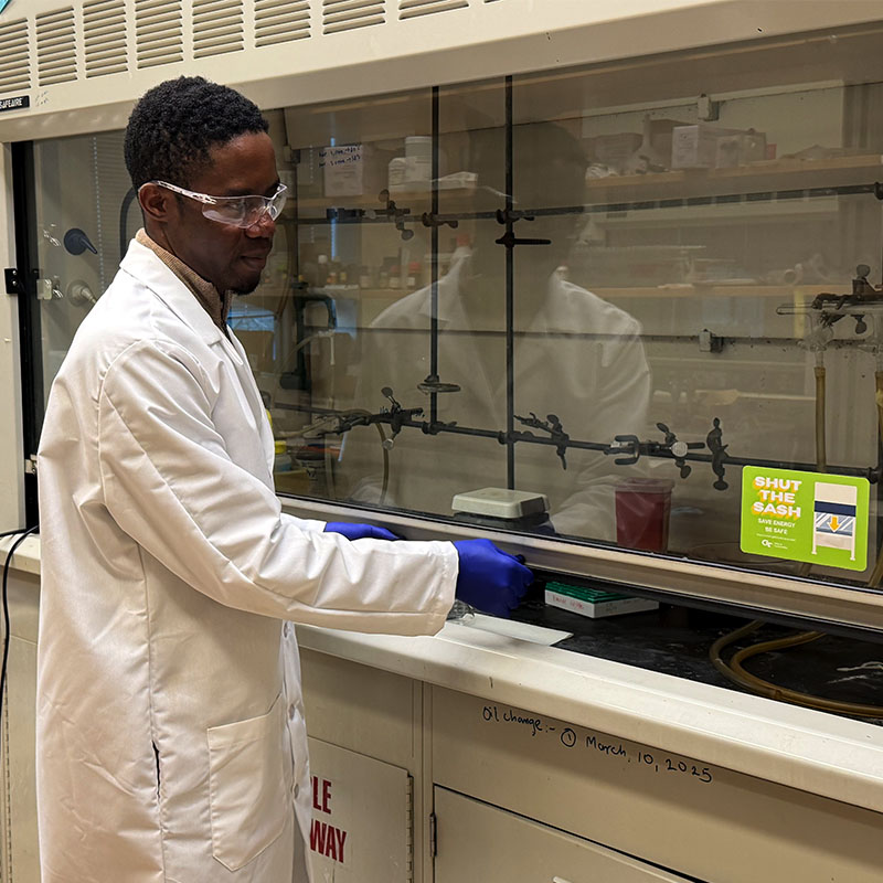 Person in a lab coat working at a chemical fume hood with gloved hands.