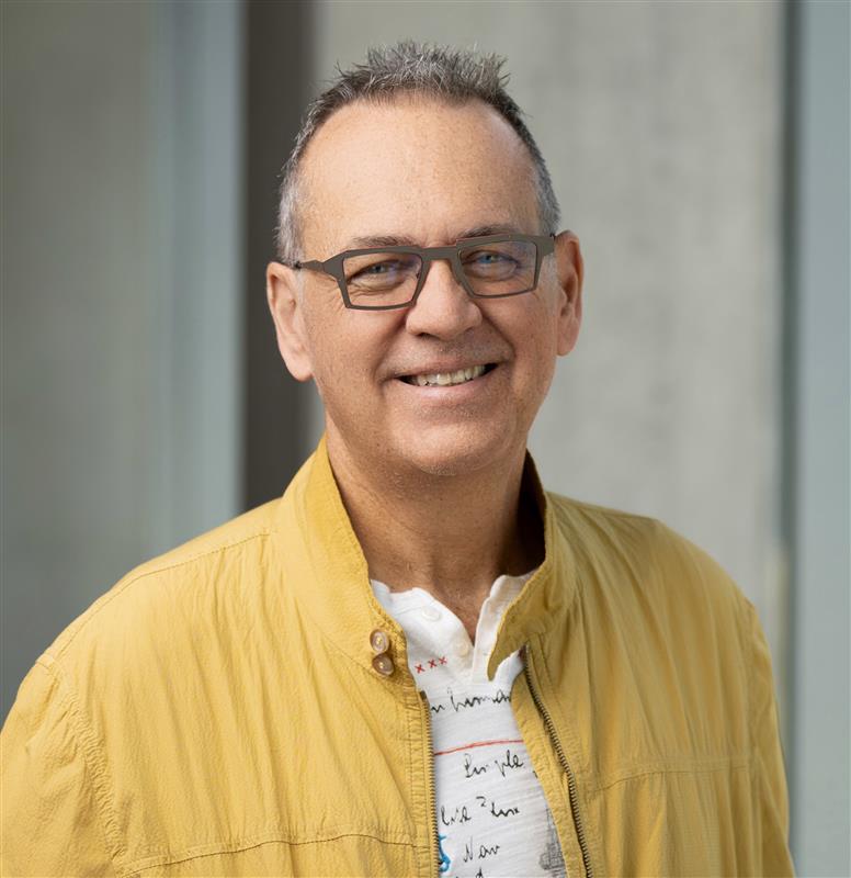 Portrait of a white man wearing glasses, a yellow jacket, and a light-colored shirt, standing in front of a softly blurred indoor background with neutral tones, looking toward the camera.