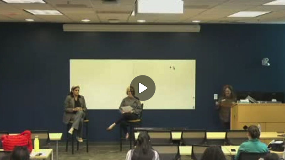 Two women sit in chairs at the front of a seminar room holding microphones. Behind them is a whiteboard and in front of them are tables and chairs with people.