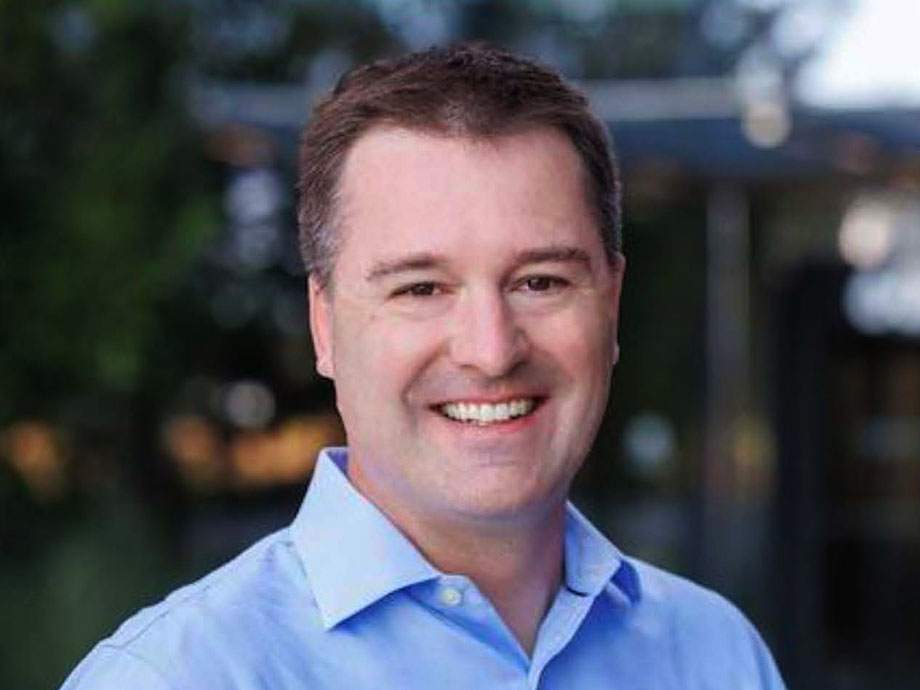 A man with short brown hair wearing a blue button-up shirt smiles for a portrait with trees and buildings in the background.