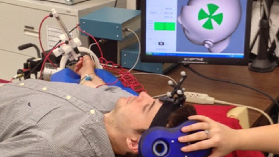 A participant lies on a lab table while researchers operate robotic and monitoring equipment connected to a computer displaying experimental data.