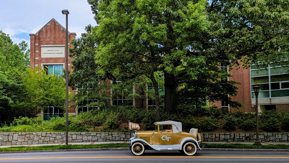  The Ramblin' Wreck, a vintage gold-and-white Georgia Tech automobile, drives along a tree-lined street in front of the Parker H. Petit Biotechnology Building on the Georgia Tech campus.