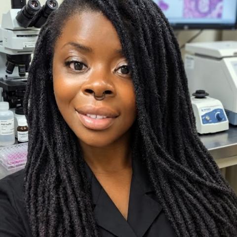 A black woman with long black braids wearing a black shirt smiles for a photo with lab equipment shown in the background.
