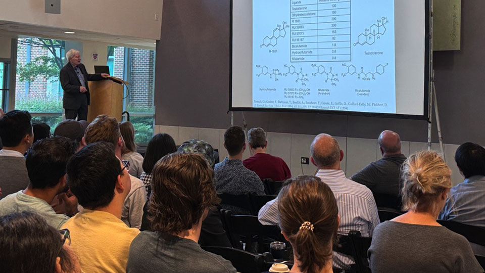 A crowd of people sit in an atrium and watch a man on a stage give a lecture while standing in front of a large projector screen.