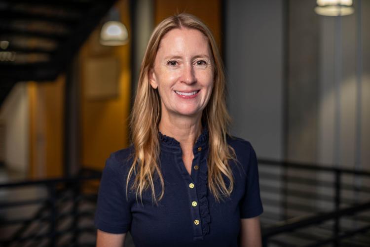A blond woman wearing a navy blue blouse smiles for a portrait with a staircase in the background